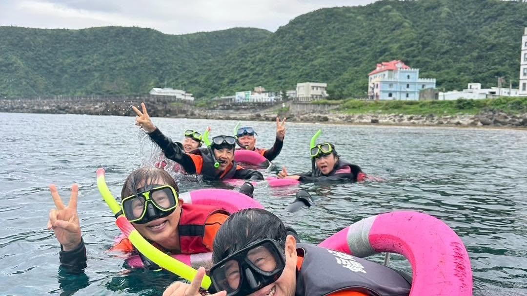浮潛中的夥伴在水面比出手勢,背後是岸邊建築