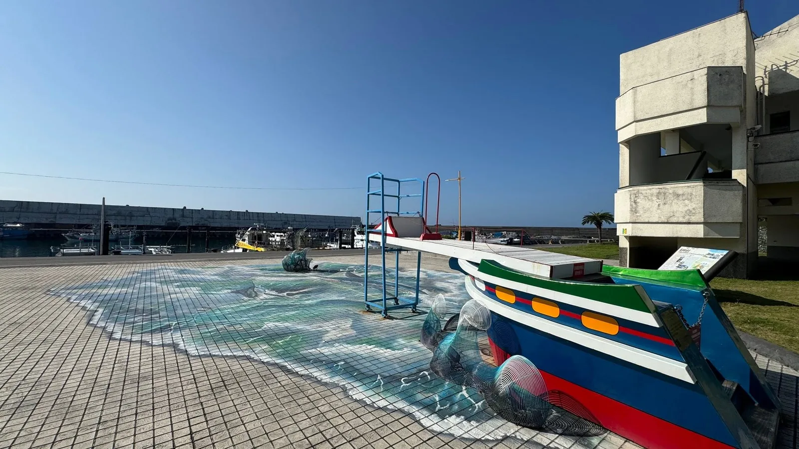 漁港旁的地面 3D 彩繪海浪與彩色漁船造景,遠方停泊著漁船