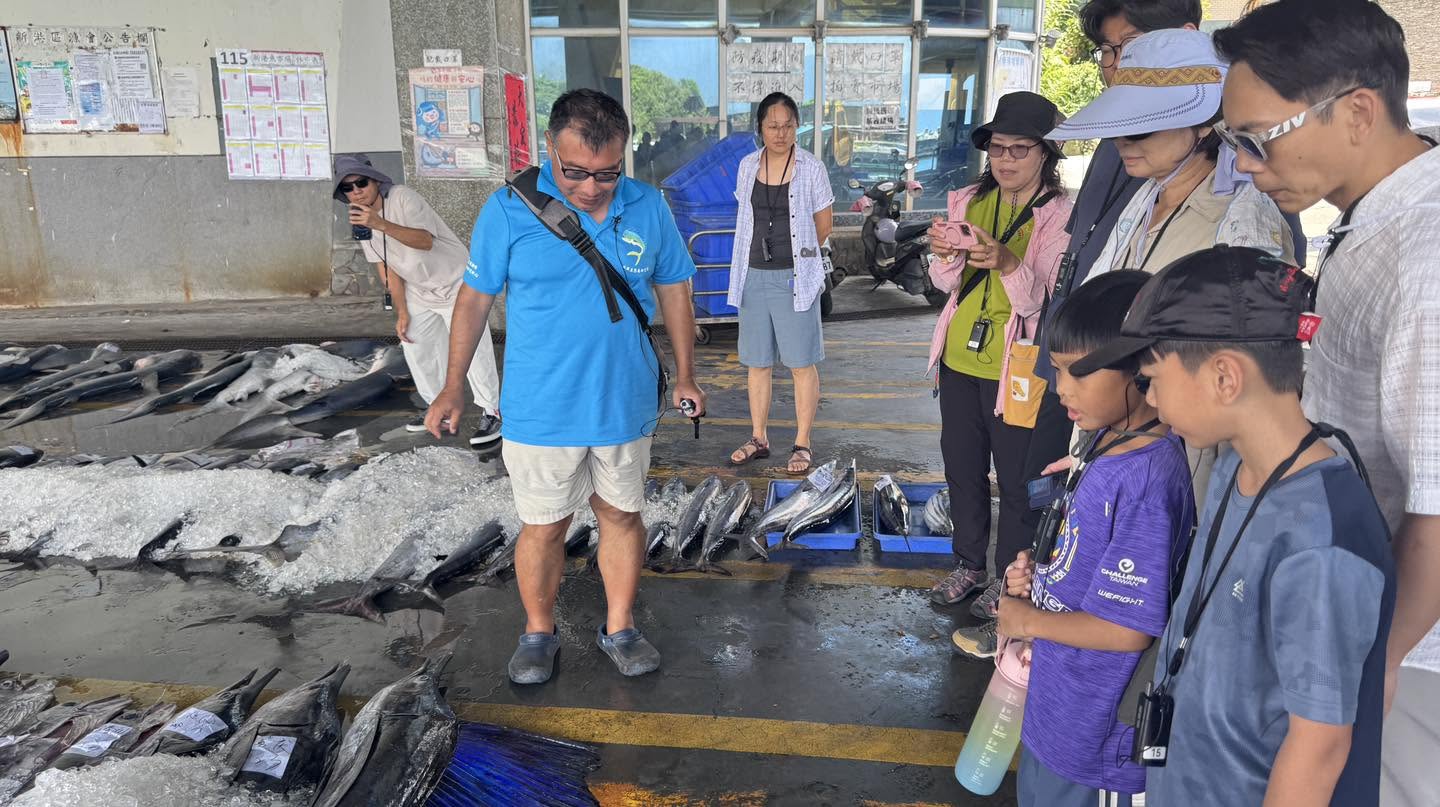 穿藍色制服的導覽人員在新港魚市場內解說,地上排列著整排上冰的旗魚,學員圍在旁邊看