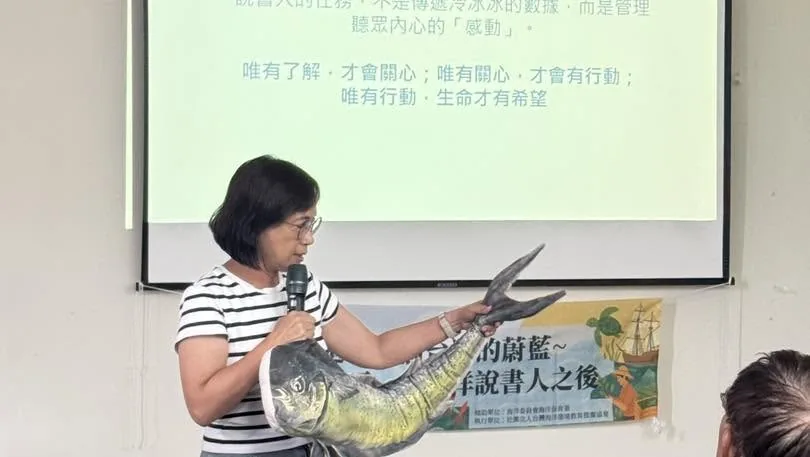 講師拿著麥克風,另一手舉起一隻鬼頭刀造型抱枕,投影片寫著「唯有了解,才會關心;唯有關心,才會行動」